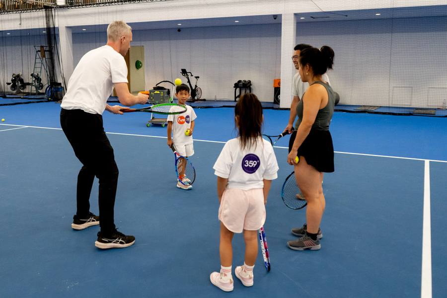 Coach geeft indoor tennisles aan kinderen en een volwassene op de blauwe banen van NTC Amstelveen — 359.tennis